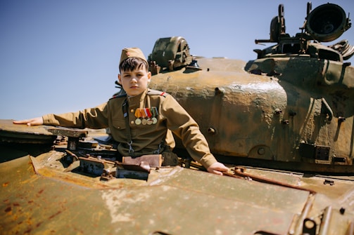 A young person wearing a military uniform with medals sits on a tank, surrounded by machinery. The sky is clear and blue in the background, conveying a sense of openness.