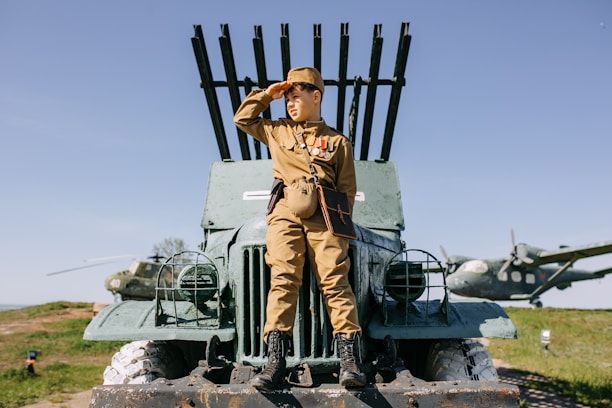 A person in vintage military attire is sitting on a green military vehicle, which has rocket launchers mounted on it. They are saluting while looking into the distance. In the background, there are two aircraft on a grassy field under a clear blue sky.