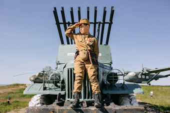 A person in vintage military attire is sitting on a green military vehicle, which has rocket launchers mounted on it. They are saluting while looking into the distance. In the background, there are two aircraft on a grassy field under a clear blue sky.