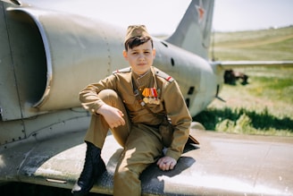 A young person dressed in a military uniform sits on the wing of a vintage aircraft. The uniform includes several medals and insignia, and the person has a calm expression. The aircraft, featuring a star emblem, is positioned in a grassy outdoor setting.