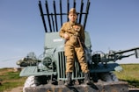 A child dressed in a military uniform sits atop an old, military vehicle. The child is wearing medals and a cap. In the background, there is a helicopter on a grassy field under a clear blue sky.