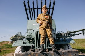 A child dressed in a military uniform sits atop an old, military vehicle. The child is wearing medals and a cap. In the background, there is a helicopter on a grassy field under a clear blue sky.
