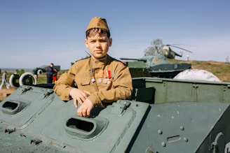 A young person dressed in a military uniform with several medals is posing on an armored vehicle. The background includes more military vehicles, a helicopter, and people casually walking or observing in what seems to be an open-air museum or exhibition setting on a clear day.