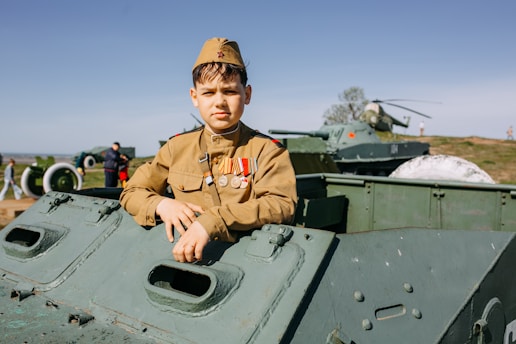 A young person dressed in a military uniform with several medals is posing on an armored vehicle. The background includes more military vehicles, a helicopter, and people casually walking or observing in what seems to be an open-air museum or exhibition setting on a clear day.