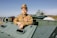 A young person dressed in a military uniform with several medals is posing on an armored vehicle. The background includes more military vehicles, a helicopter, and people casually walking or observing in what seems to be an open-air museum or exhibition setting on a clear day.