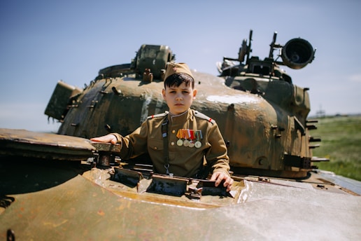 A child dressed in a military uniform with several medals is standing inside a tank. The scene takes place outdoors on a clear day with green grass visible in the background.