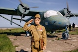 A young person dressed in a vintage military uniform, complete with medals, salutes in front of a weathered, older model airplane with significant chipping paint. The backdrop includes a grassy field and a clear blue sky, adding to a historical ambiance.