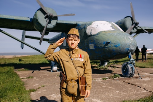 A young person dressed in a vintage military uniform, complete with medals, salutes in front of a weathered, older model airplane with significant chipping paint. The backdrop includes a grassy field and a clear blue sky, adding to a historical ambiance.