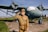 A young person dressed in a vintage military uniform, complete with medals, salutes in front of a weathered, older model airplane with significant chipping paint. The backdrop includes a grassy field and a clear blue sky, adding to a historical ambiance.