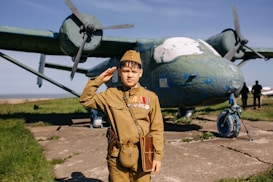 A young person dressed in a vintage military uniform, complete with medals, salutes in front of a weathered, older model airplane with significant chipping paint. The backdrop includes a grassy field and a clear blue sky, adding to a historical ambiance.