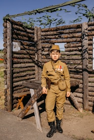 A young person dressed in a vintage military uniform stands in front of a rustic structure made from logs. The uniform includes a hat, medals, and a pouch, and the person appears to be posing for the photograph. The background structure resembles a log cabin or fort, with greenery and a net above.