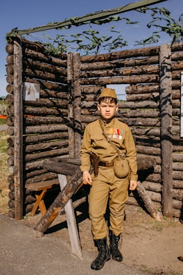 A young person dressed in a vintage military uniform stands in front of a rustic structure made from logs. The uniform includes a hat, medals, and a pouch, and the person appears to be posing for the photograph. The background structure resembles a log cabin or fort, with greenery and a net above.