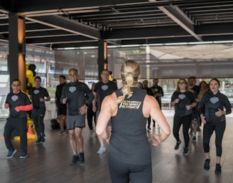 A group of individuals in athletic wear participate in a fitness class inside a spacious, well-lit room. The instructor, facing away, leads the group with energy and focus. The participants appear engaged and attentive, following along with the instructor's movements.