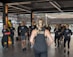 A group of individuals in athletic wear participate in a fitness class inside a spacious, well-lit room. The instructor, facing away, leads the group with energy and focus. The participants appear engaged and attentive, following along with the instructor's movements.