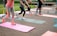 People are exercising on yoga mats laid out on a brick pavement. They are performing a group workout outdoors, with several bottles of brightly colored sports drinks placed beside the mats. The participants are dressed in athletic clothing, some in leggings and others in shorts.