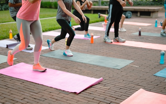 People are exercising on yoga mats laid out on a brick pavement. They are performing a group workout outdoors, with several bottles of brightly colored sports drinks placed beside the mats. The participants are dressed in athletic clothing, some in leggings and others in shorts.