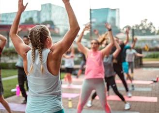 A group of people is participating in an outdoor fitness class on a paved area with yoga mats laid out. In the foreground, a person with braided hair, wearing a sleeveless top and leggings, raises their arms. Other participants are dressed in sportswear, energetically following the movements of the instructor.