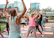 A group of people is participating in an outdoor fitness class on a paved area with yoga mats laid out. In the foreground, a person with braided hair, wearing a sleeveless top and leggings, raises their arms. Other participants are dressed in sportswear, energetically following the movements of the instructor.