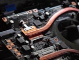 A close-up view of a circuit board featuring copper heat pipes, various electronic components, connectors, and chips. The copper elements stand out against the dark, intricate background of the circuit board.