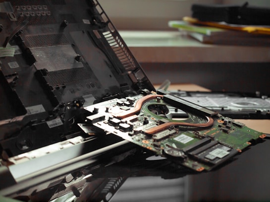 A disassembled laptop sits on a desk with its internal components exposed, including the motherboard and cooling system. Various other pieces, like the battery and outer casing, are also visible around the laptop. The workspace appears cluttered, and a stack of folders or papers can be seen in the background.