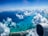 Aerial view of an airplane wing and engine against a backdrop of white clouds and vibrant blue ocean waters. The coastline is visible in the lower part of the image, with a mix of developed areas and natural landscapes. Fluffy clouds are scattered throughout the sky, adding depth to the scene.