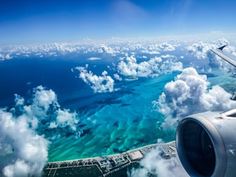 Aerial view of an airplane wing and engine against a backdrop of white clouds and vibrant blue ocean waters. The coastline is visible in the lower part of the image, with a mix of developed areas and natural landscapes. Fluffy clouds are scattered throughout the sky, adding depth to the scene.