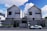 Two modern, symmetrical, double-story houses with minimalistic architectural features. The houses have white walls with dark windows and a geometric patterned facade at the front. A sleek sports car is parked on the street in front of the house. The sky is blue with scattered clouds.