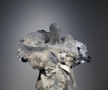 A mannequin is adorned with an elaborate and intricate dress featuring voluminous floral decorations and ribbons. The design includes shimmering elements and textured layers, creating a luxurious and artistic appearance.