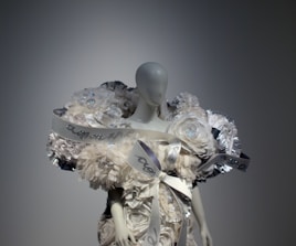 A mannequin is adorned with an elaborate and intricate dress featuring voluminous floral decorations and ribbons. The design includes shimmering elements and textured layers, creating a luxurious and artistic appearance.