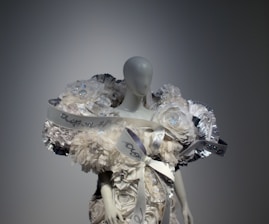 A mannequin is adorned with an elaborate and intricate dress featuring voluminous floral decorations and ribbons. The design includes shimmering elements and textured layers, creating a luxurious and artistic appearance.