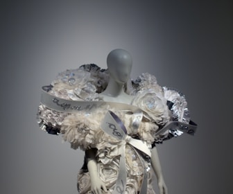 A mannequin is adorned with an elaborate and intricate dress featuring voluminous floral decorations and ribbons. The design includes shimmering elements and textured layers, creating a luxurious and artistic appearance.