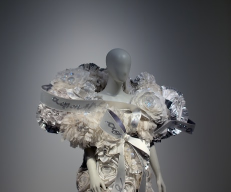 A mannequin is adorned with an elaborate and intricate dress featuring voluminous floral decorations and ribbons. The design includes shimmering elements and textured layers, creating a luxurious and artistic appearance.