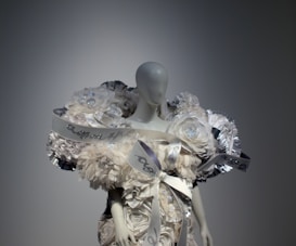 A mannequin is adorned with an elaborate and intricate dress featuring voluminous floral decorations and ribbons. The design includes shimmering elements and textured layers, creating a luxurious and artistic appearance.