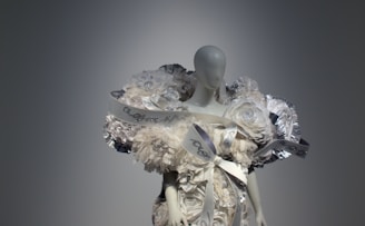 A mannequin dressed in an elaborate outfit made of layered, voluminous fabric resembling white flowers and ribbons. The design includes metallic accents and intricate details, creating an avant-garde, sculptural appearance.