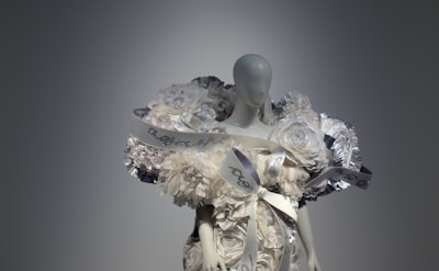 A mannequin dressed in an elaborate outfit made of layered, voluminous fabric resembling white flowers and ribbons. The design includes metallic accents and intricate details, creating an avant-garde, sculptural appearance.
