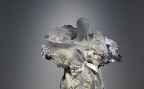 A mannequin dressed in an elaborate outfit made of layered, voluminous fabric resembling white flowers and ribbons. The design includes metallic accents and intricate details, creating an avant-garde, sculptural appearance.