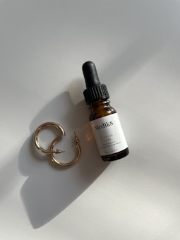 A brown dropper bottle labeled 'Medik8 C-Tetra Luxe' is positioned next to a pair of gold hoop earrings on a light surface. The arrangement casts soft shadows, and the light reflects on the metallic surface of the earrings.