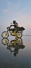 A motorcyclist wearing protective gear, including a helmet and gloves, rides a motorcycle across a reflective, wet surface. The scene captures a striking reflection of both the rider and the bike on the water-like surface, under a soft evening or dawn sky with subtle pastel tones.