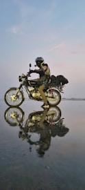 A motorcyclist wearing protective gear, including a helmet and gloves, rides a motorcycle across a reflective, wet surface. The scene captures a striking reflection of both the rider and the bike on the water-like surface, under a soft evening or dawn sky with subtle pastel tones.