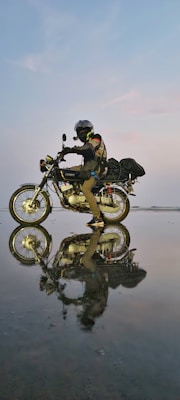 A motorcyclist wearing protective gear, including a helmet and gloves, rides a motorcycle across a reflective, wet surface. The scene captures a striking reflection of both the rider and the bike on the water-like surface, under a soft evening or dawn sky with subtle pastel tones.
