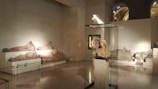 A museum gallery displays several classical sculptures. In the foreground, a bust is enclosed in a glass case. On the walls, there are reclining figures, likely sarcophagi, strategically lit to highlight their contours. The setting is spacious, with high ceilings and soft lighting, contributing to a serene ambiance.