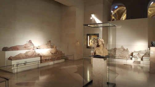 A museum gallery displays several classical sculptures. In the foreground, a bust is enclosed in a glass case. On the walls, there are reclining figures, likely sarcophagi, strategically lit to highlight their contours. The setting is spacious, with high ceilings and soft lighting, contributing to a serene ambiance.