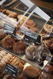 A variety of baked goods are displayed in a glass case. There are round cakes topped with cocoa powder, crescent-shaped pastries with a glazed finish, and other assorted baked treats. Small black signs with white text indicate the names and prices of the items, written in a foreign language.