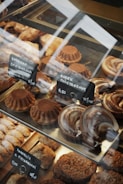 A variety of baked goods are displayed in a glass case. There are round cakes topped with cocoa powder, crescent-shaped pastries with a glazed finish, and other assorted baked treats. Small black signs with white text indicate the names and prices of the items, written in a foreign language.