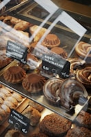 A variety of baked goods are displayed in a glass case. There are round cakes topped with cocoa powder, crescent-shaped pastries with a glazed finish, and other assorted baked treats. Small black signs with white text indicate the names and prices of the items, written in a foreign language.