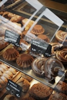 A variety of baked goods are displayed in a glass case. There are round cakes topped with cocoa powder, crescent-shaped pastries with a glazed finish, and other assorted baked treats. Small black signs with white text indicate the names and prices of the items, written in a foreign language.