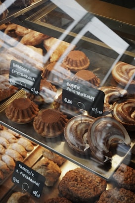 A variety of baked goods are displayed in a glass case. There are round cakes topped with cocoa powder, crescent-shaped pastries with a glazed finish, and other assorted baked treats. Small black signs with white text indicate the names and prices of the items, written in a foreign language.
