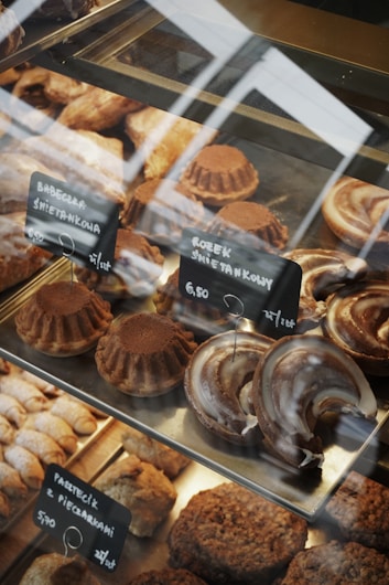 A variety of baked goods are displayed in a glass case. There are round cakes topped with cocoa powder, crescent-shaped pastries with a glazed finish, and other assorted baked treats. Small black signs with white text indicate the names and prices of the items, written in a foreign language.