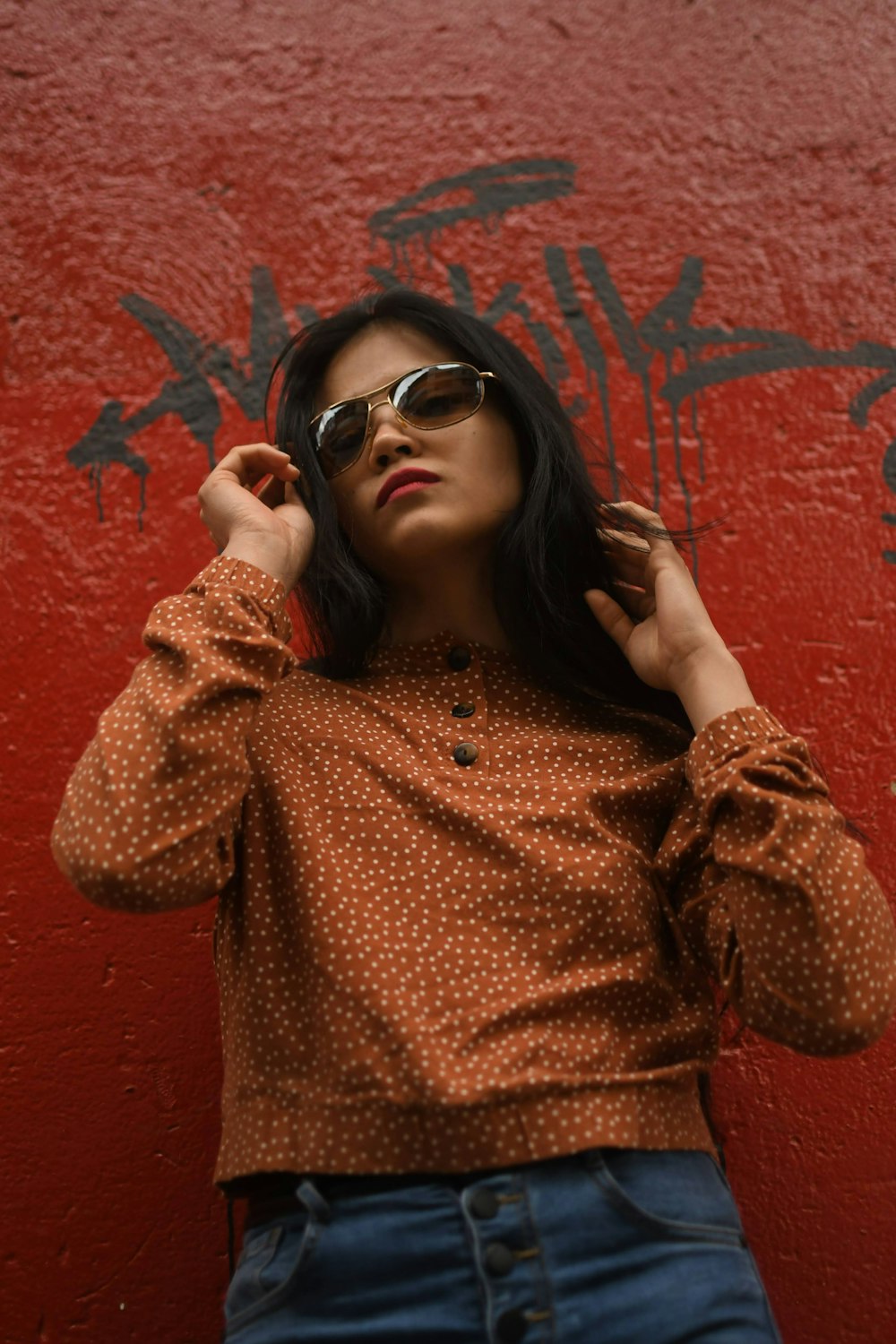 a woman in a brown shirt and sunglasses leaning against a red wall