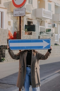 A person stands on a street holding up a large blue right arrow sign, with the sign covering their face. Behind the person, there is a traffic post with Italian text and a no-entry symbol. The background includes a rough stone wall and residential buildings with balconies.